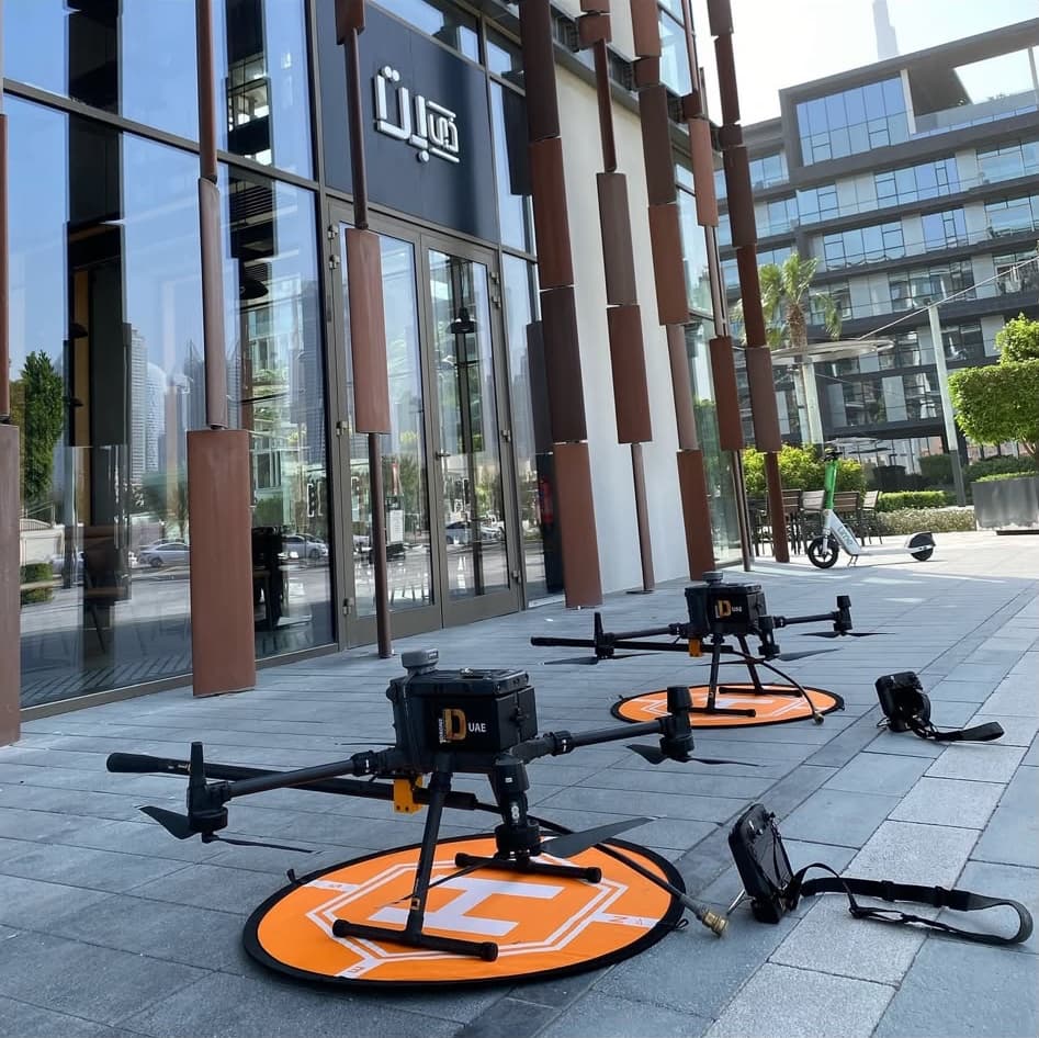 Two working drones on orange landing pads on the pavement outside a glass office building